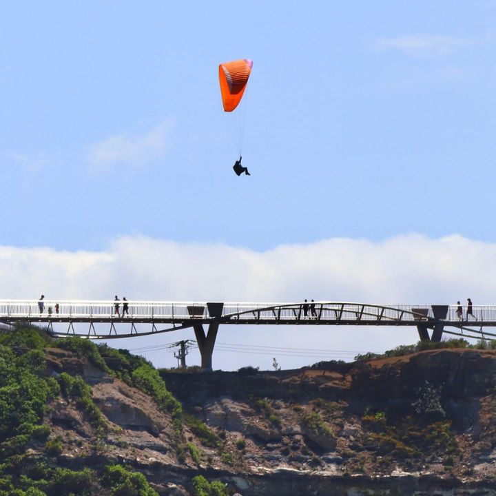 A paraglider flys above a steel arch walkway